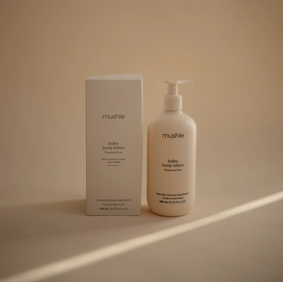 A bottle of mushie baby body lotion with a pump next to its white cardboard box, set against a warm beige background with soft diagonal shadows.