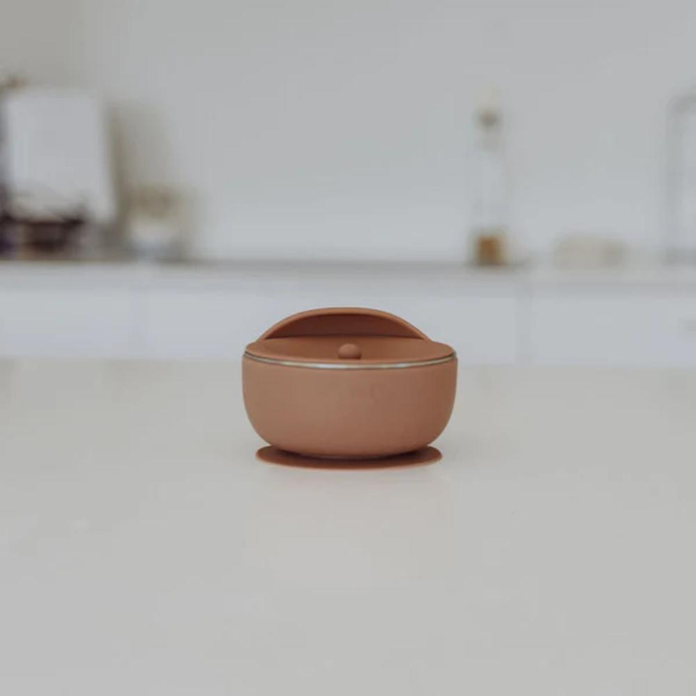 A small terracotta-colored silicone lidded container with a rounded dome lid and a tiny central knob, sitting on a light gray surface with a blurred, minimalist background.