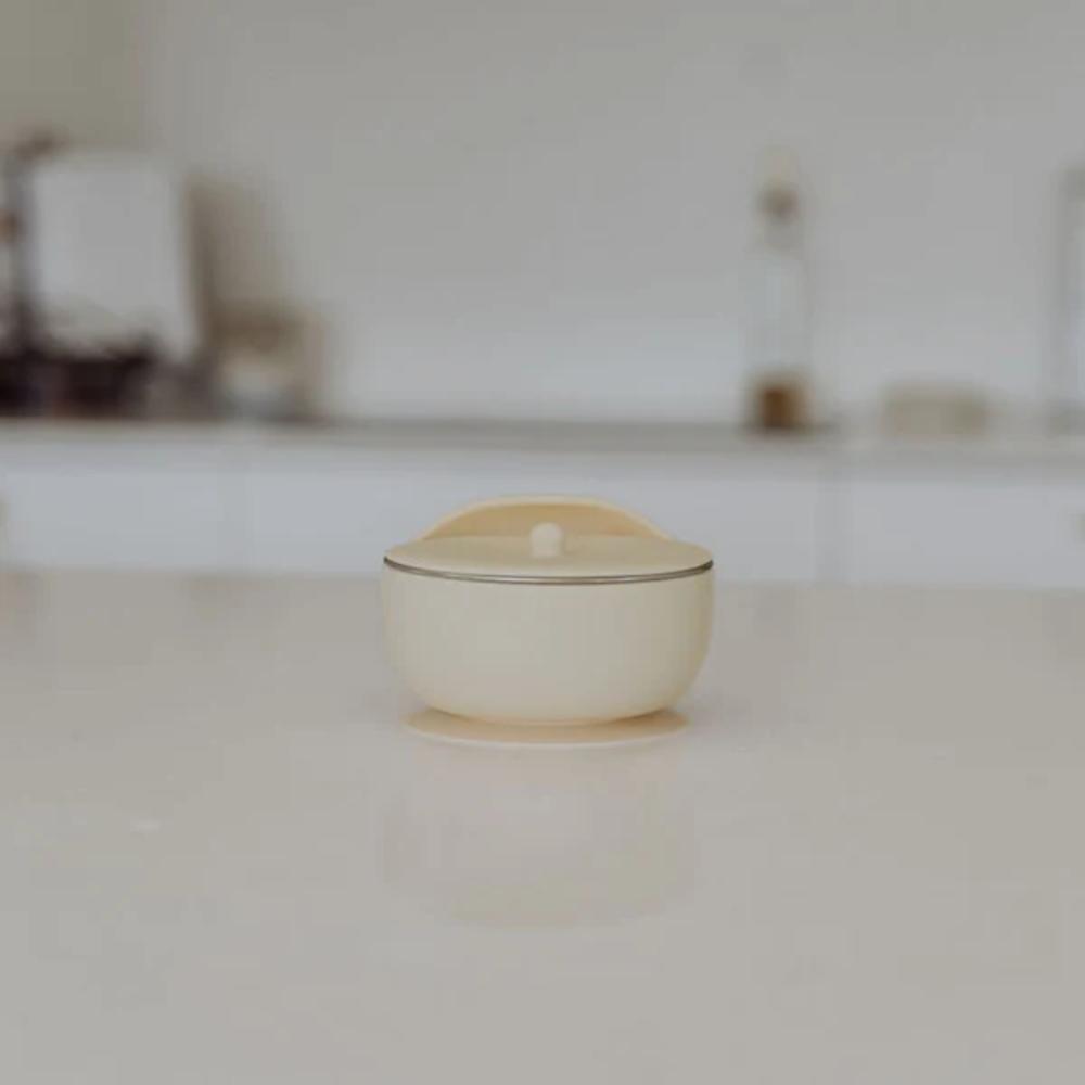 A small cream-colored silicone container with a matching lid and stainless steel bowl set on a light-colored countertop with a blurred kitchen background.