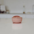A small dusty pink silicone lidded container with stainless steel bowl set on a clean white surface with a blurred kitchen background.