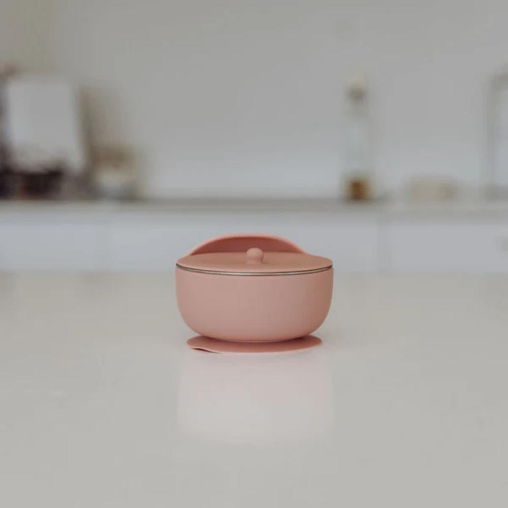 A small dusty pink silicone lidded container with stainless steel bowl set on a clean white surface with a blurred kitchen background.
