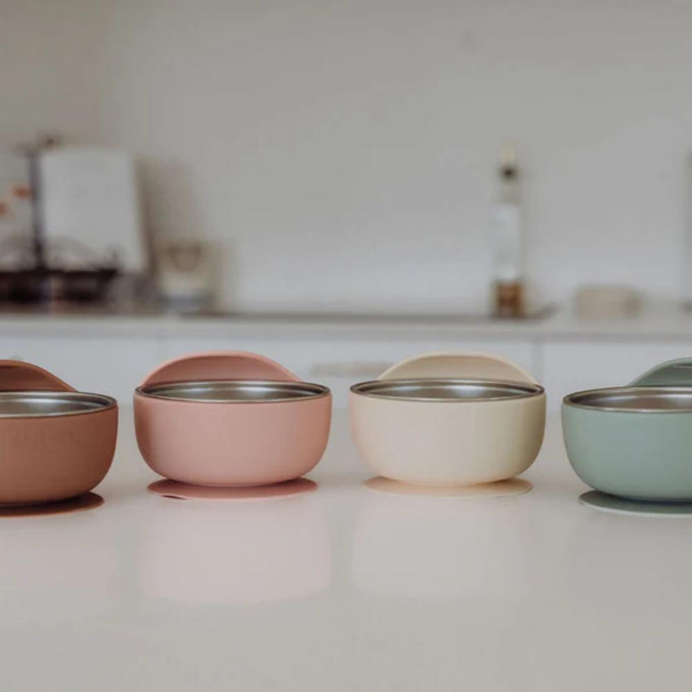 Four pastel-colored silicone bowls with stainless steel interiors lined up on a white countertop in a softly blurred kitchen setting.