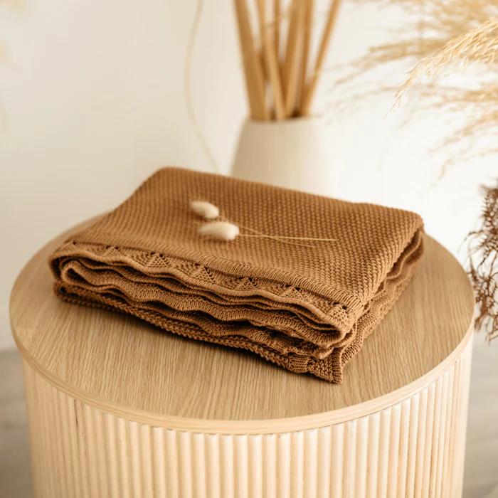 A folded brown textured knit blanket with a scalloped edge, displayed on a round wooden pedestal with dried grasses in the background.