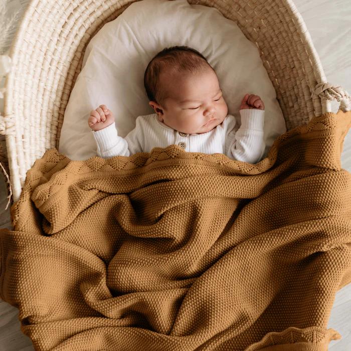 A sleeping newborn baby in a white ribbed long-sleeve outfit, nestled inside a woven natural fiber bassinet and wrapped in a textured brown blanket.