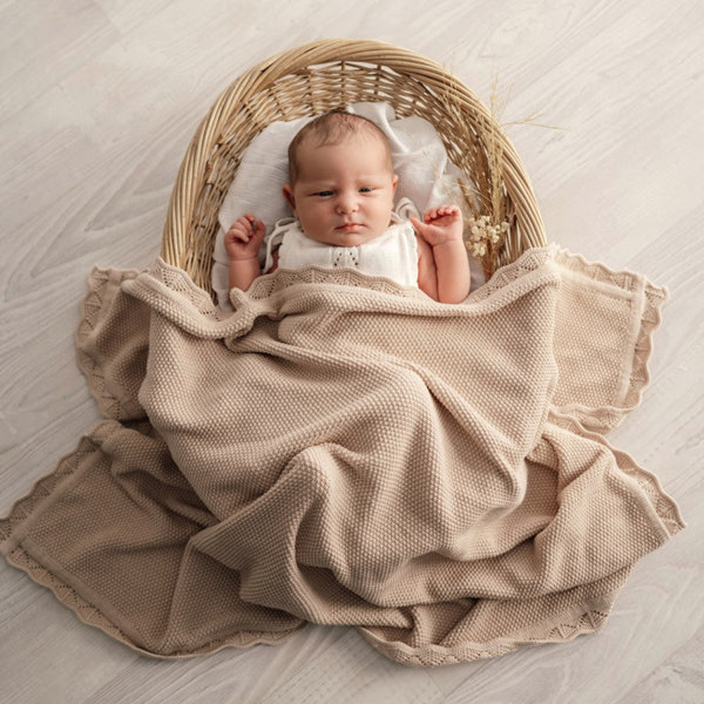 A baby wrapped in a beige textured blanket, lying inside a woven wicker basket on a light wood floor.