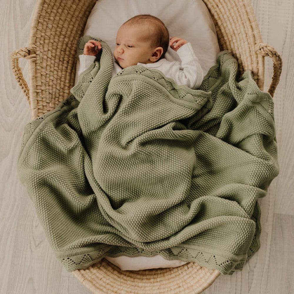 A newborn baby wrapped in an olive green textured knit blanket, lying inside a woven straw Moses basket on a light wood floor.