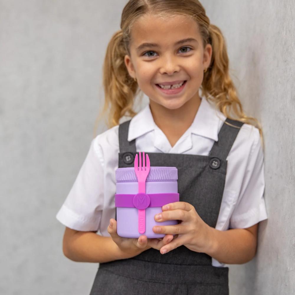 A young girl with blonde pigtails smiles while holding a purple lunch container with a pink silicone fork, wearing a white collared shirt and dark gray overalls against a textured gray wall.