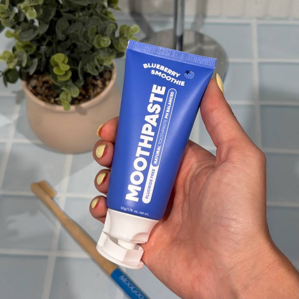 Hand holding a blue tube of 'Blueberry Smoothie' toothpaste with a plant and toothbrush in the background.