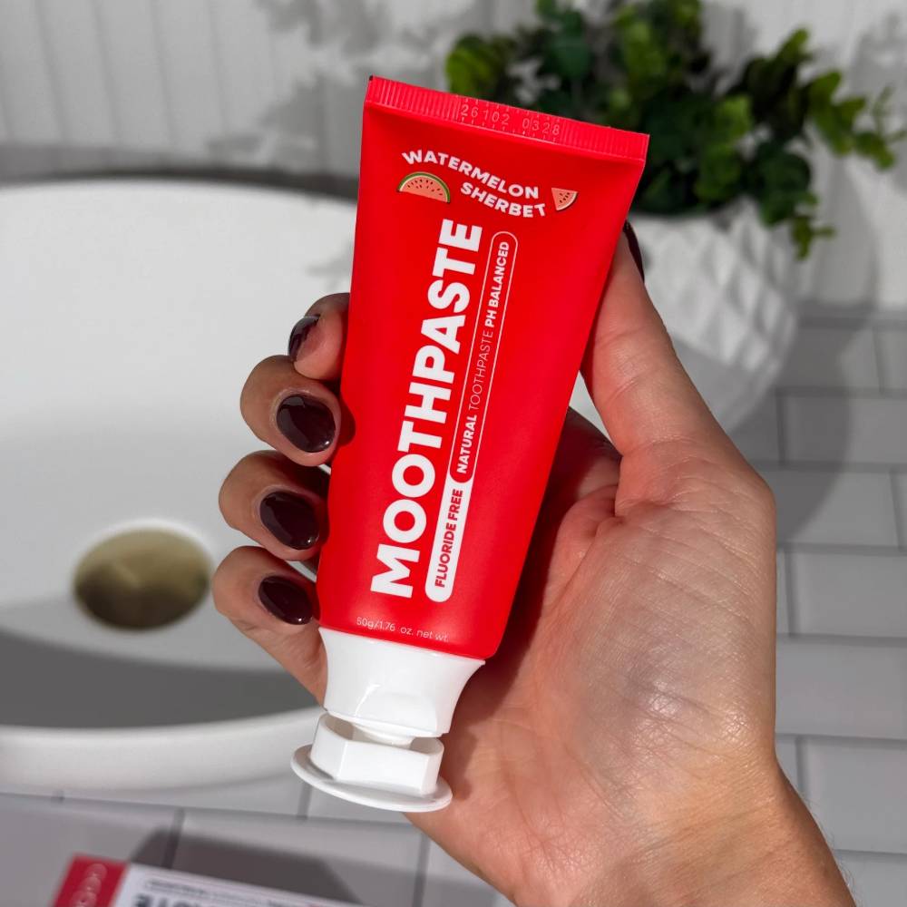Hand holding a red tube of 'Moothpaste' with a blurred bathroom background