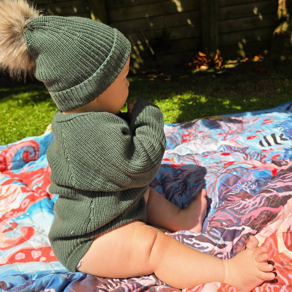 Child wearing a green merino wool knit outfit and matching hat with pom-pom sitting on a colorful blanket outdoors.