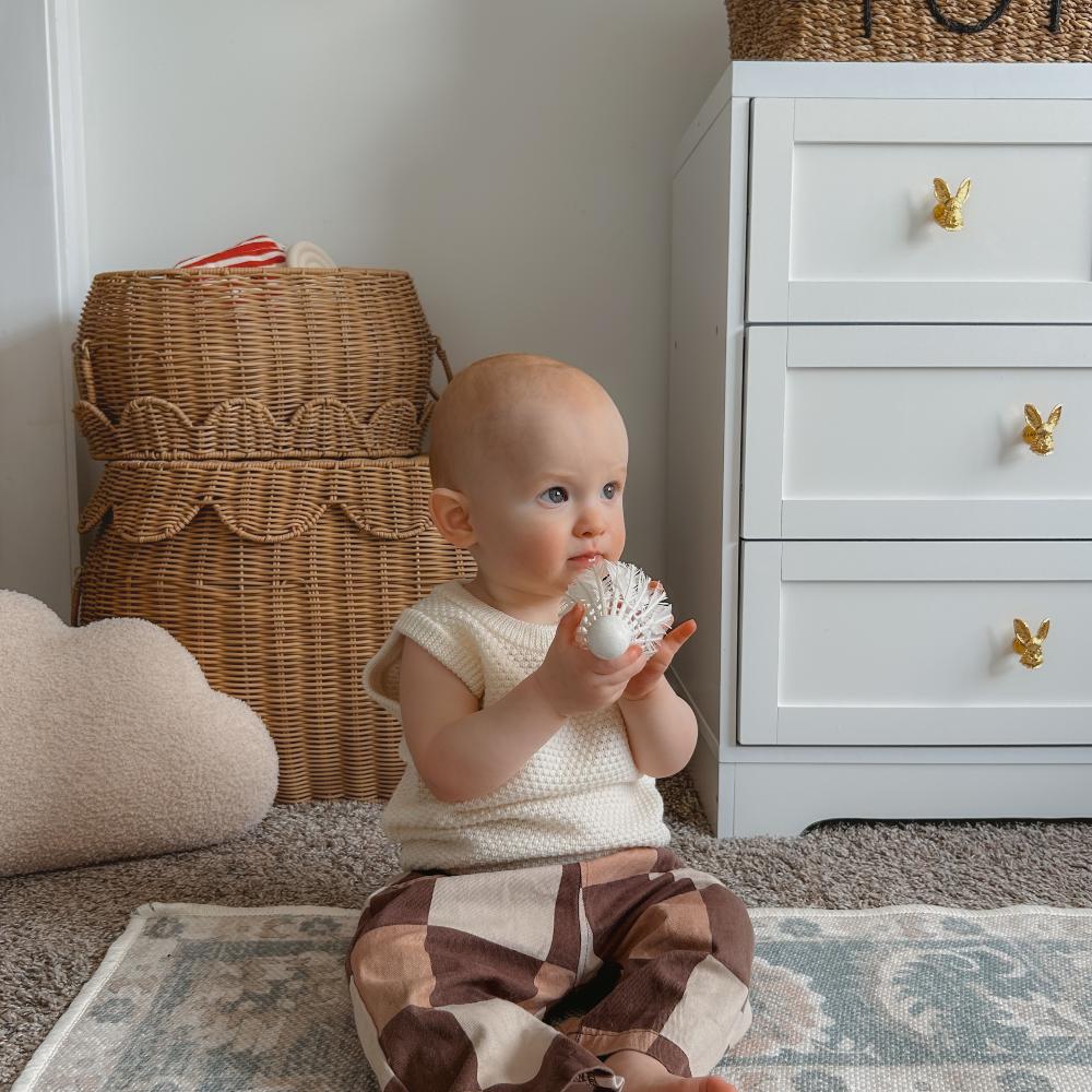 Baby sitting on a rug holding a white object in a room with a wicker basket and white dresser wearing a cream knit vest and brown patterned pants