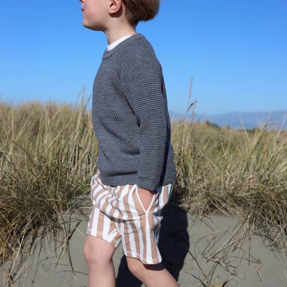 Child in a grey woolen knitted sweater and striped shorts standing on a sandy beach with grasses.