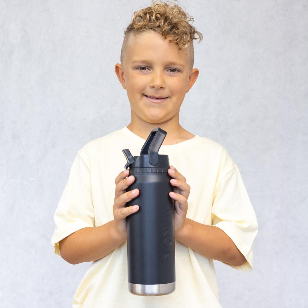 A young boy with curly light brown hair wearing a cream-colored t-shirt holds a matte black insulated water bottle with a stainless steel base against a textured gray background.