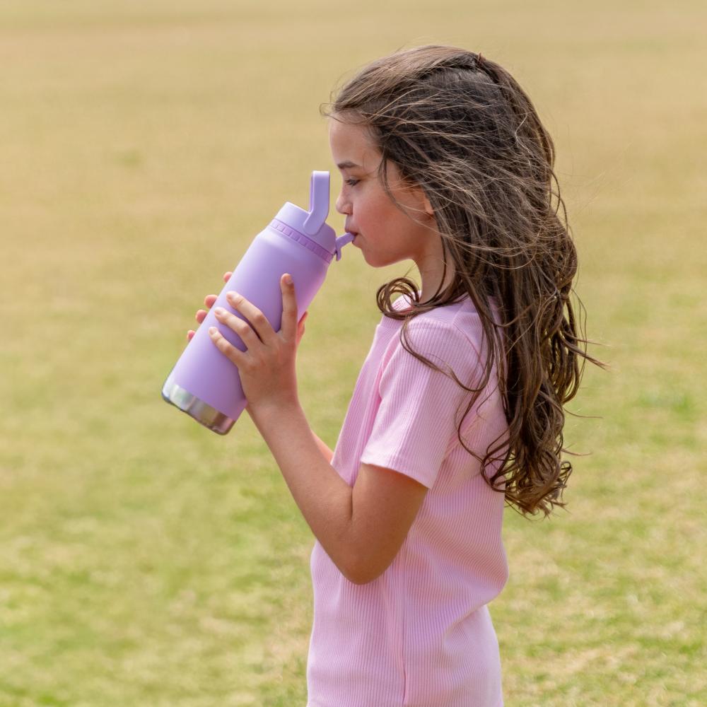 A young girl with long brown hair is shown in profile drinking from a lavender purple insulated water bottle with a matching purple lid, set against a soft-focus grassy field background.