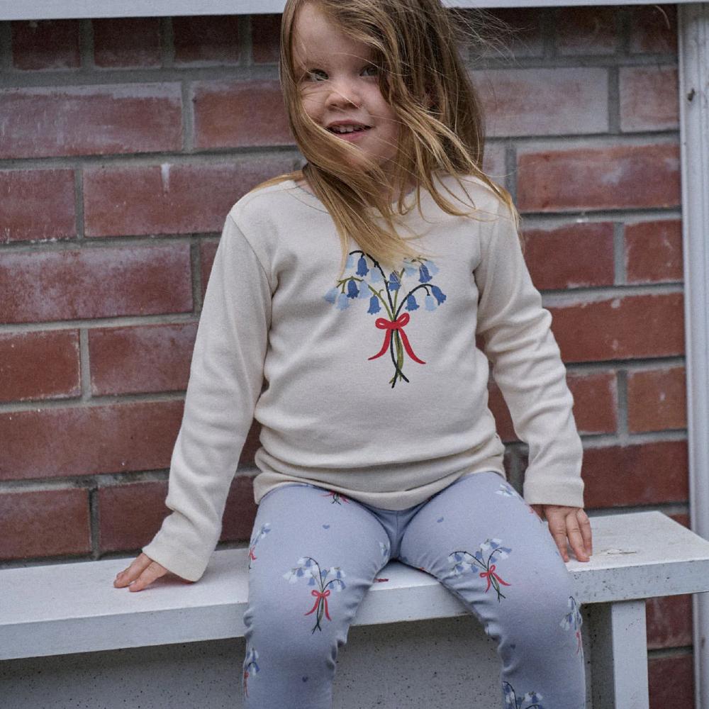 Child wearing a cream coloured sweater with a bluebell floral design and matching blue pants, sitting on a bench against a brick wall.