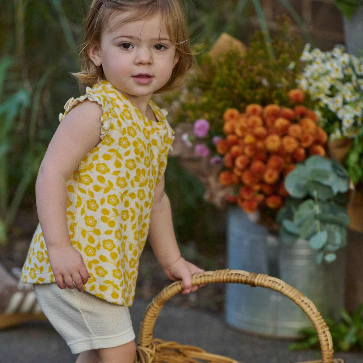 A young child wearing a yellow sleeveless top with a yellow floral print and white shorts, standing outdoors near a wicker basket and flowers.