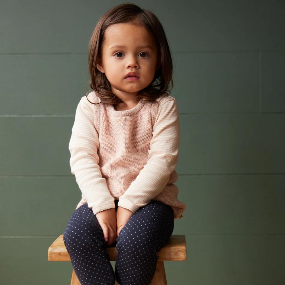 Young child sitting on a wooden stool against a green wall wearing a merino wool pink knitted vest and blue dotted leggings