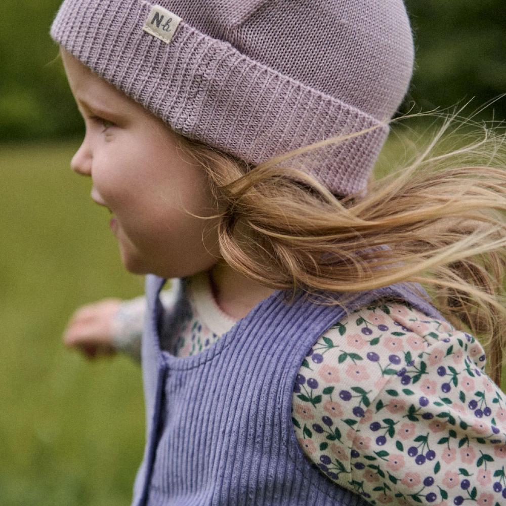 Child wearing a lilac coloured knit hat and purple corded outfit with floral pattern in an outdoor setting