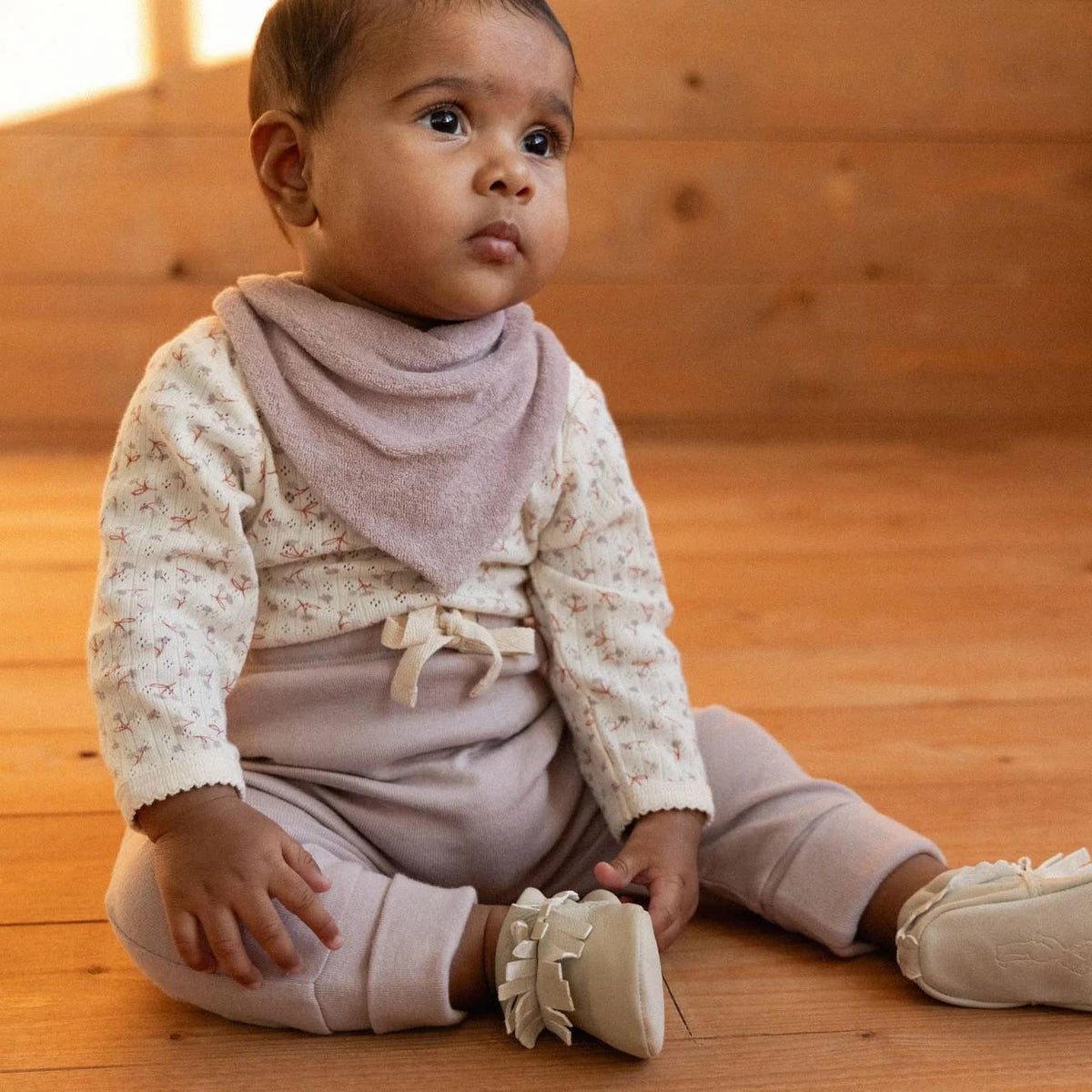 A baby sitting on a wooden floor wearing a cream floral long-sleeve romper with a dusty mauve bib and matching pants.