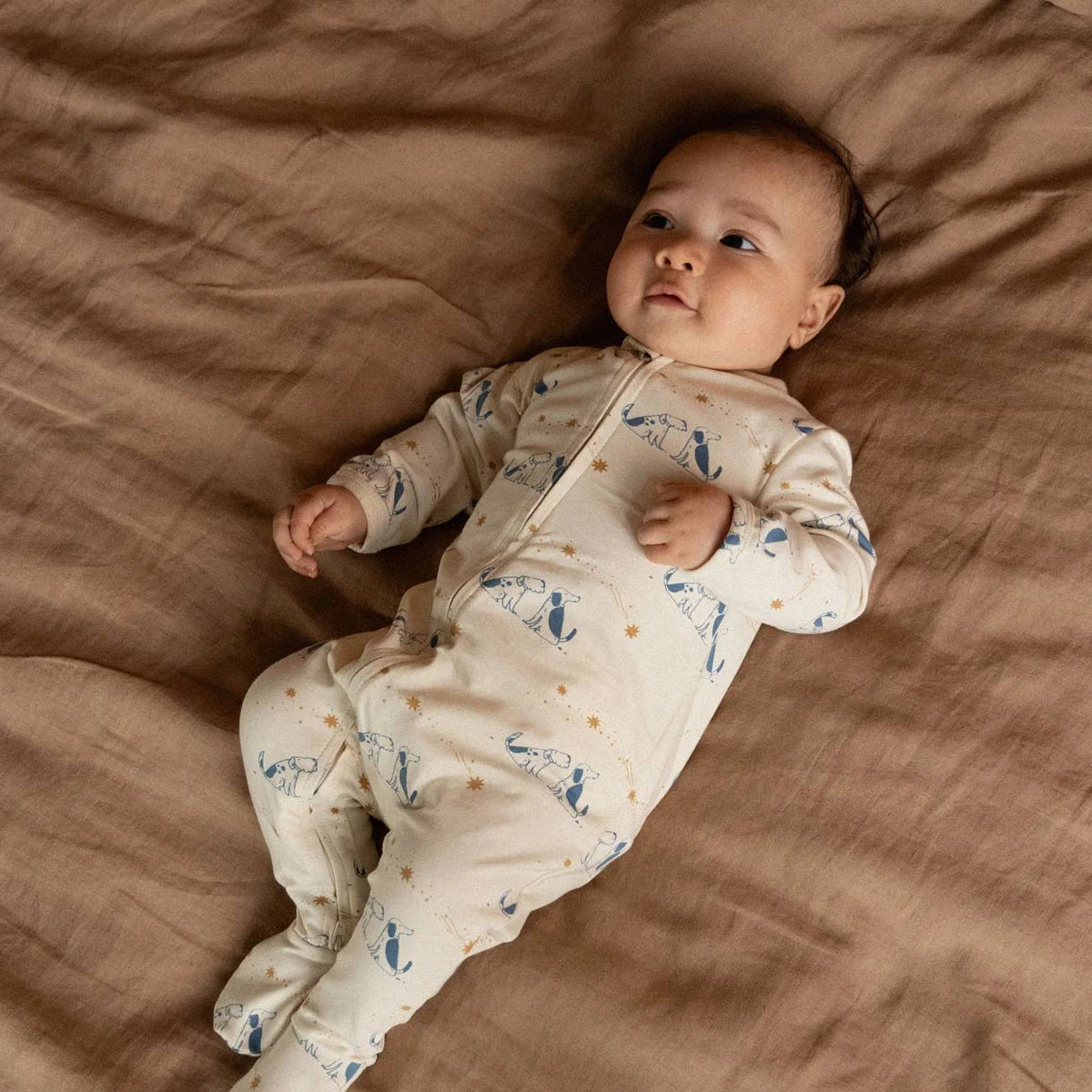 A baby lying on their back on a brown linen blanket, wearing a cream-colored footed sleeper with a blue and yellow dog print.