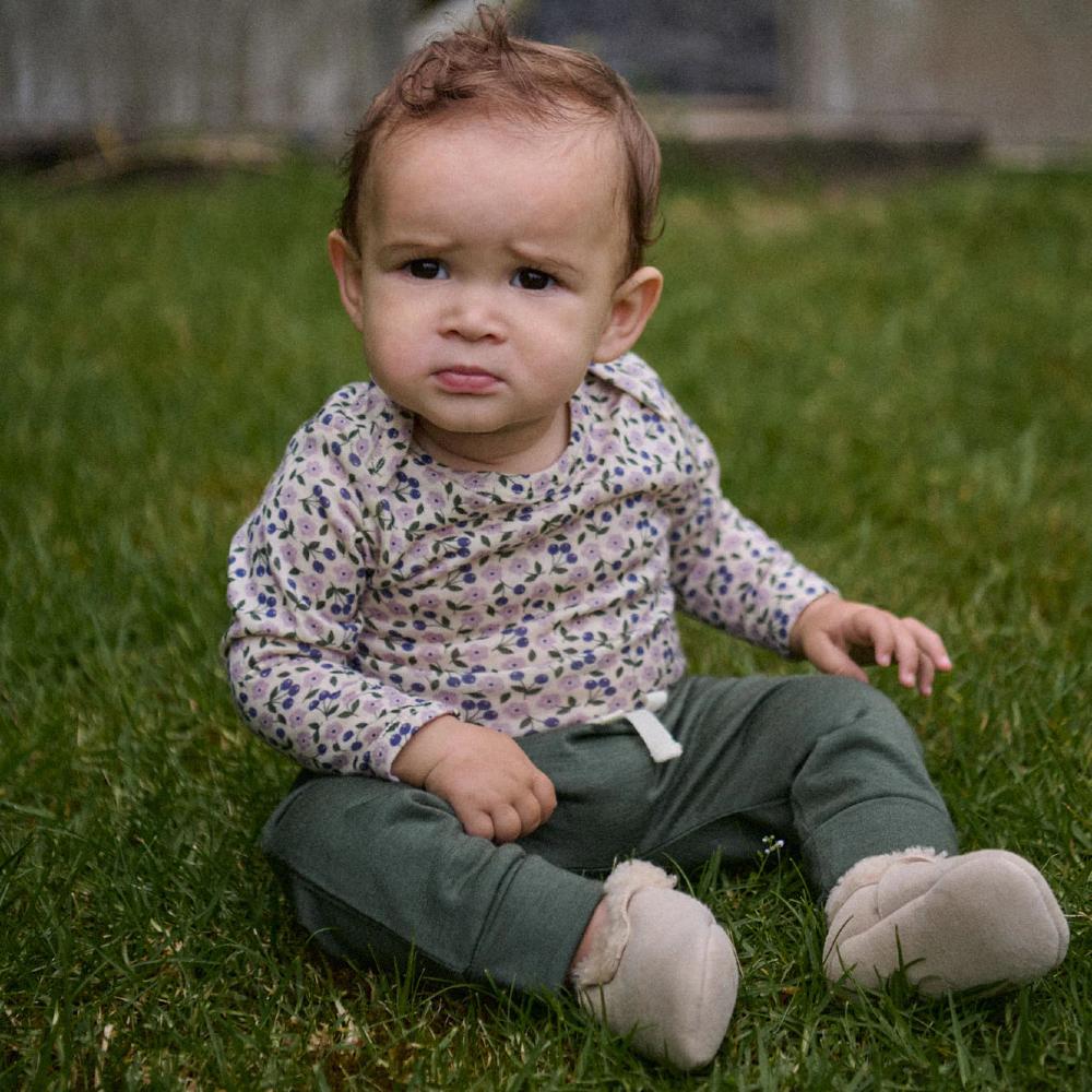 Baby sitting on grass wearing a floral bodysuit and forest green pants.