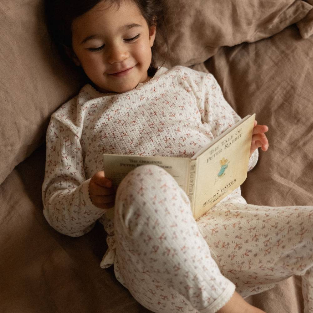 A young child lying on a brown bed, smiling while reading a book. The child is wearing a cream-colored long-sleeve pajama top and matching pants with a small red floral pattern.