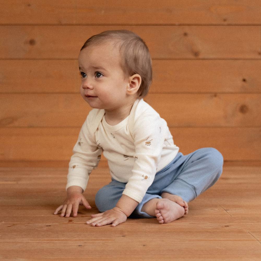 A baby sitting cross-legged on a wooden floor against a wooden plank wall, wearing a cream long-sleeve bodysuit with small motifs and light blue pants.