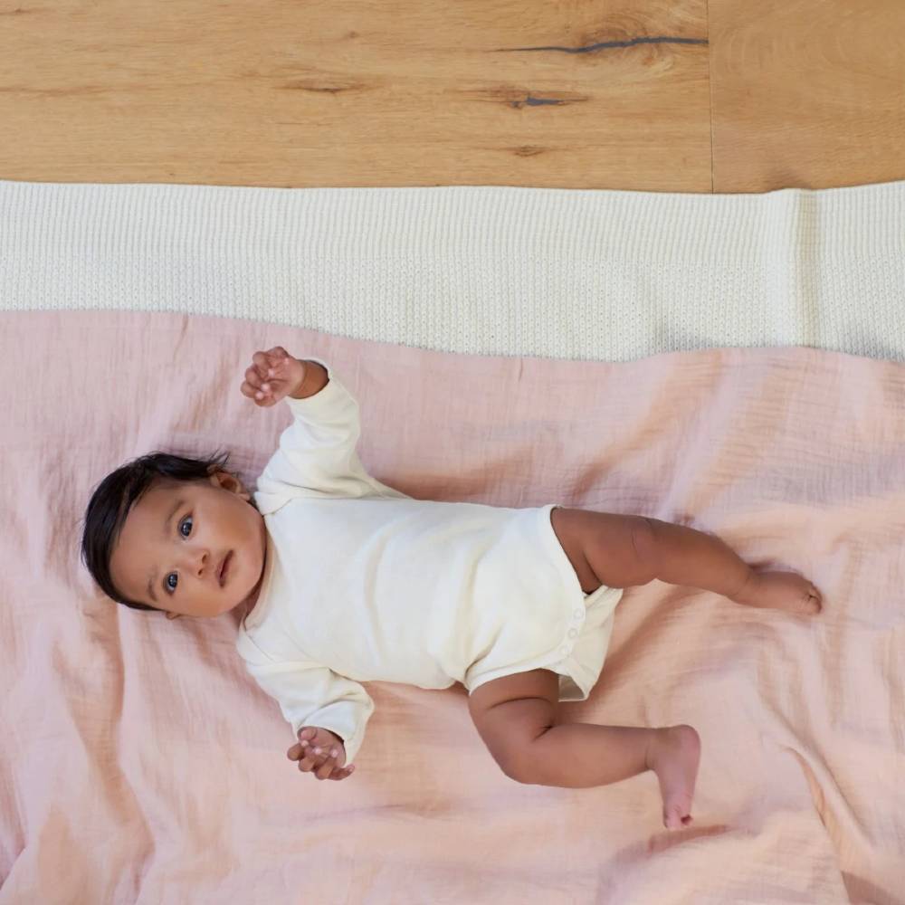 Baby lying on a pink blanket with a wooden floor in the background wearing a cream cotton baby bodysuit with long sleeves and envelope neck on a white background