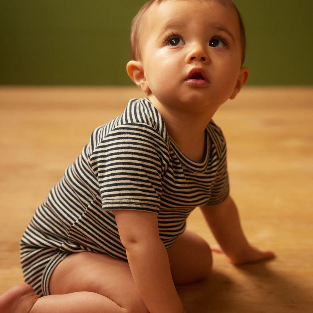 Baby in a navy and white nature baby striped onesie sitting on a wooden floor with a green wall background