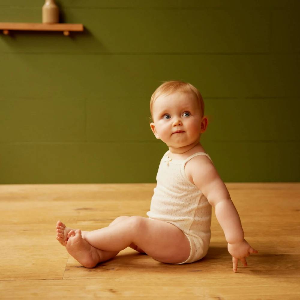 A baby sitting on the floor wearing a singlet bodysuit in a natural colour. 