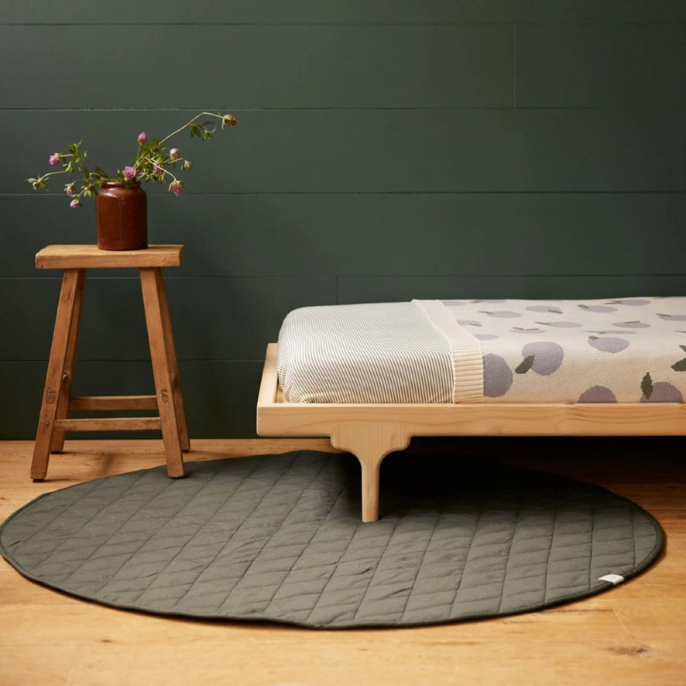 Round gray quilted play mat on a wooden floor with a bed and stool against a dark green wall.