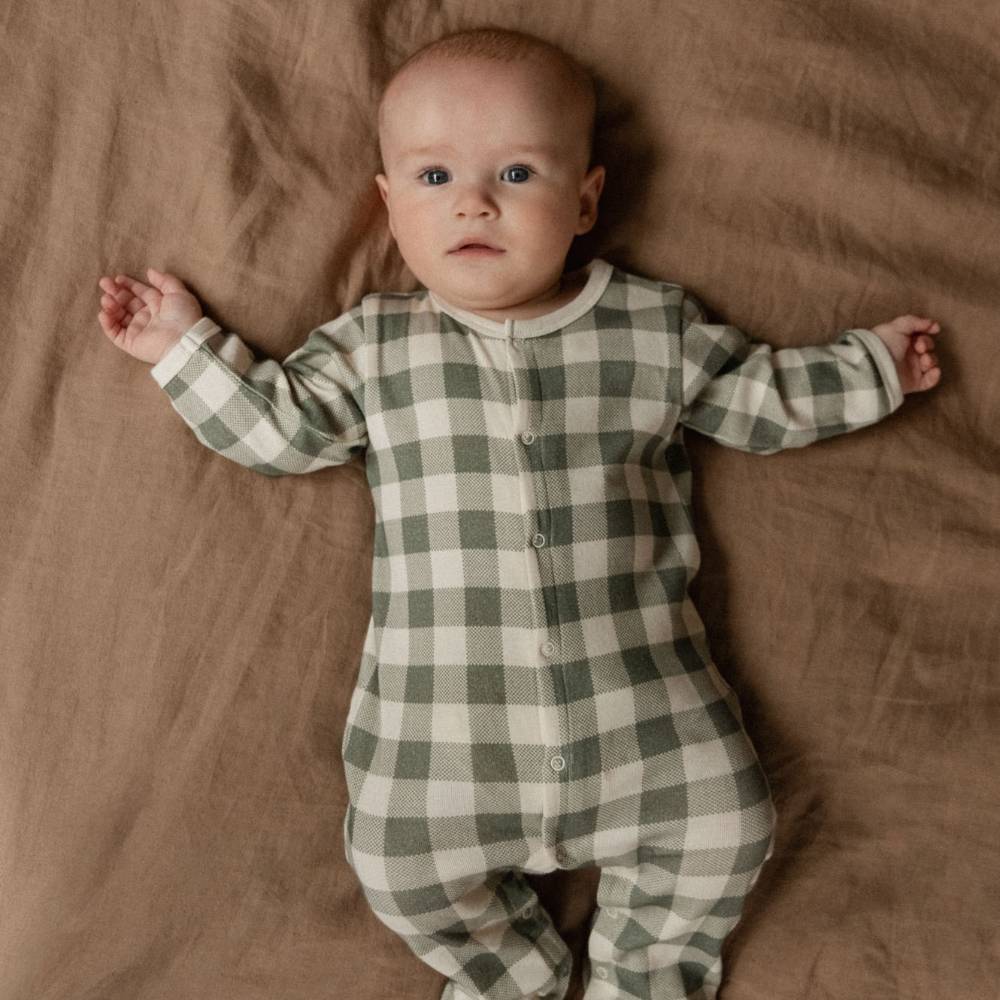 A baby lying on a brown textured blanket, wearing a long-sleeve button-front footed sleeper in an olive green and cream gingham check pattern.