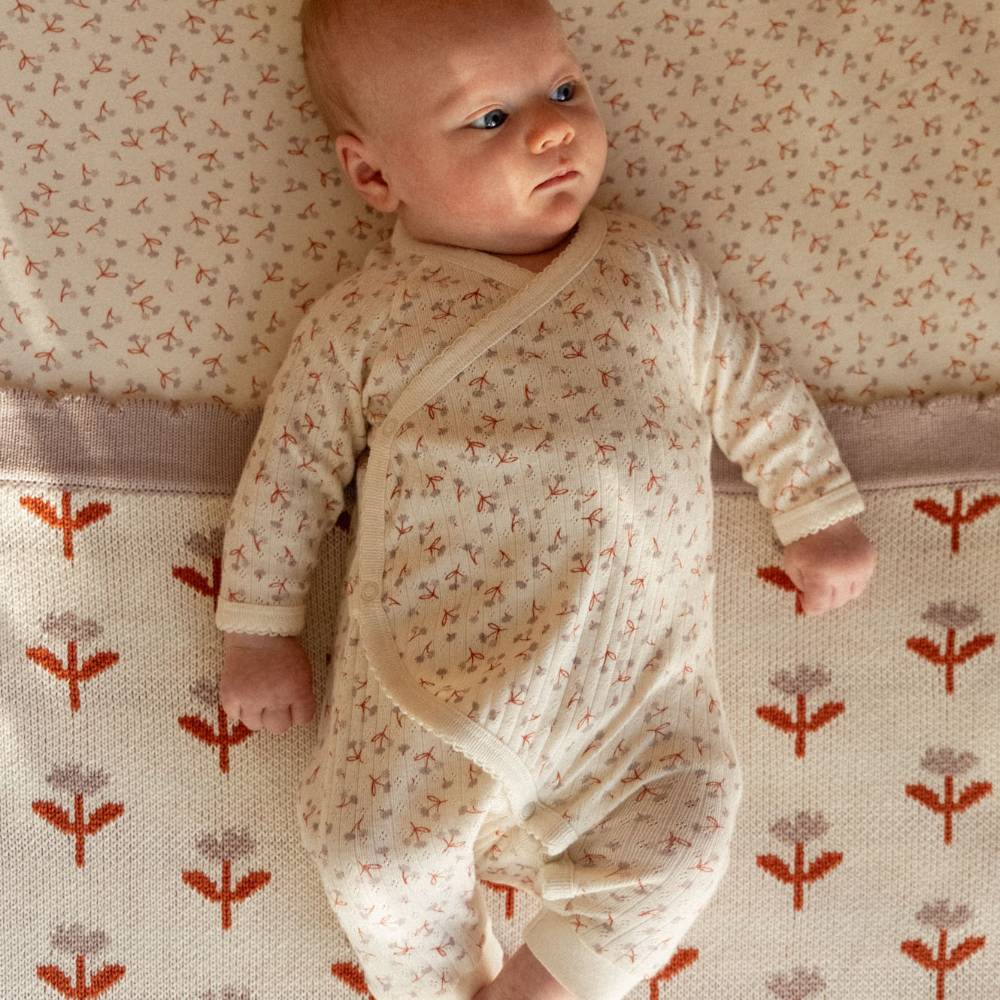 A baby lying on a patterned blanket wearing a cream-colored long-sleeve wrap onesie with a small floral print.