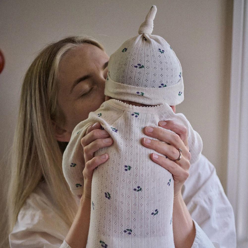 Woman holding a baby wearing a cotton pointelle longsleeve bodysuit with winter berry patterns, wearing a matching white knit hat.