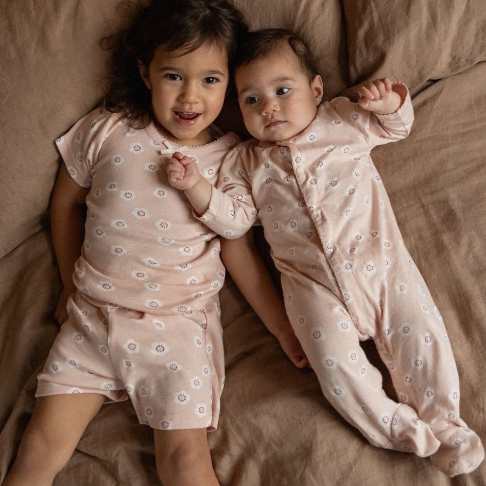 A toddler and an infant lying side by side on a brown fabric surface, wearing matching pale pink short-sleeve pajamas with a small white floral print.