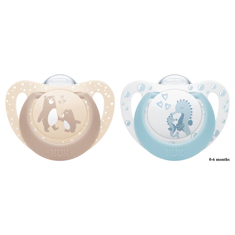 Two NUK baby pacifiers on a white background, one in beige with a penguin design and one in light blue with a dinosaur design. Small text on the right reads '0-6 months'.
