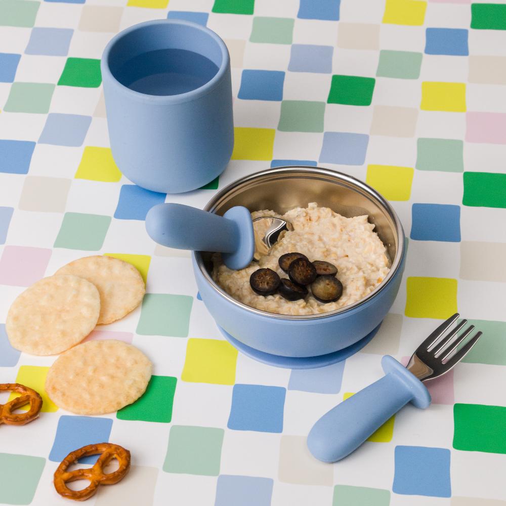 Toddlers silicone and stainless steel tableware set with blue cup, bowl, and utensils on a checkered tablecloth.