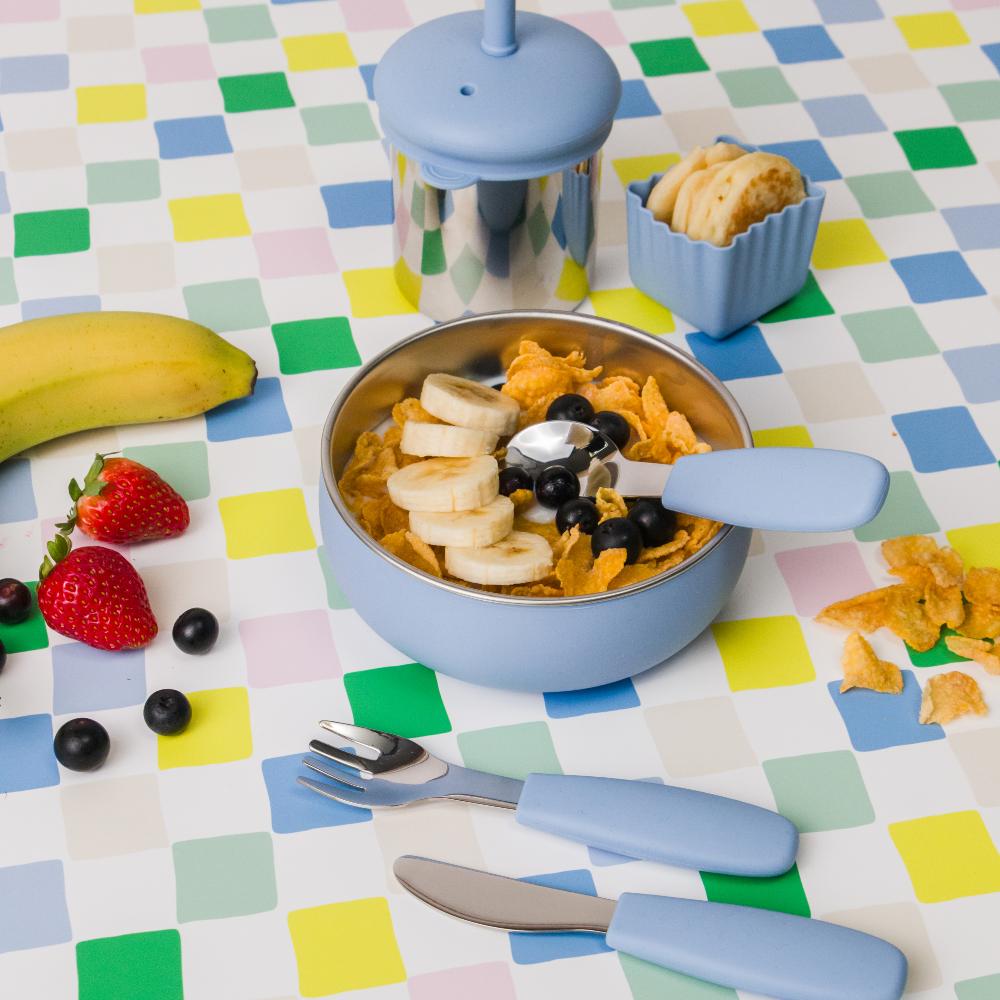 Toddler stainless steel meal set with bowl, fork, and knife on a colorful checkered tablecloth.