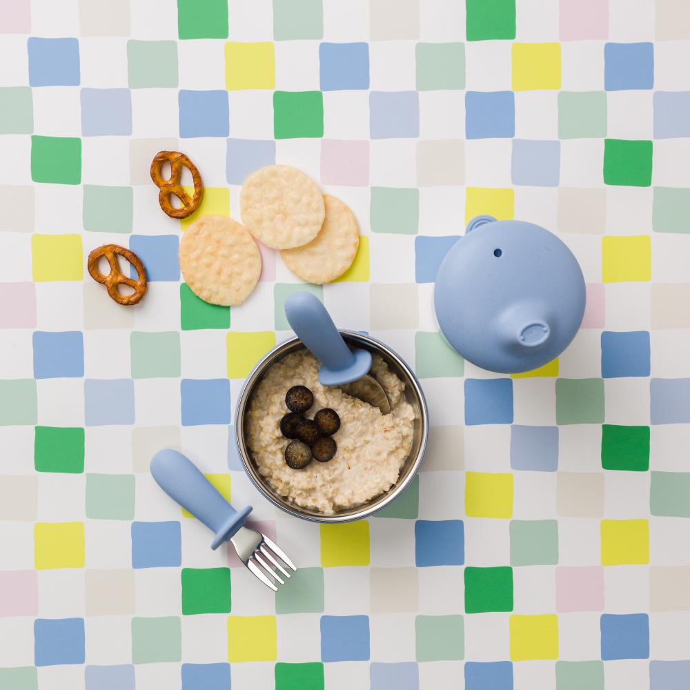 Toddler's table setting with food, spoon, fork and cup on a colorful checkered tablecloth