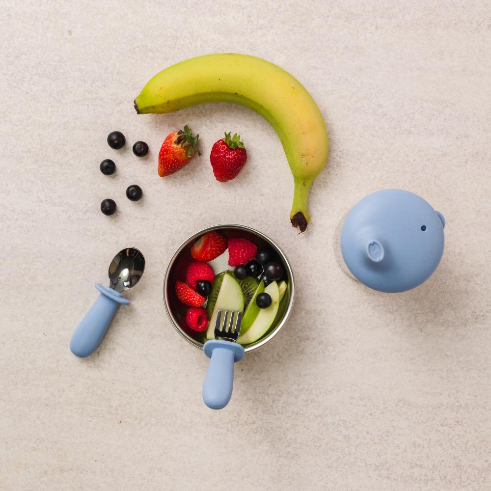 Nestling stainless steel baby bowl with fork and spoon and sippy cup on table with banana, strawberries and blueberries.