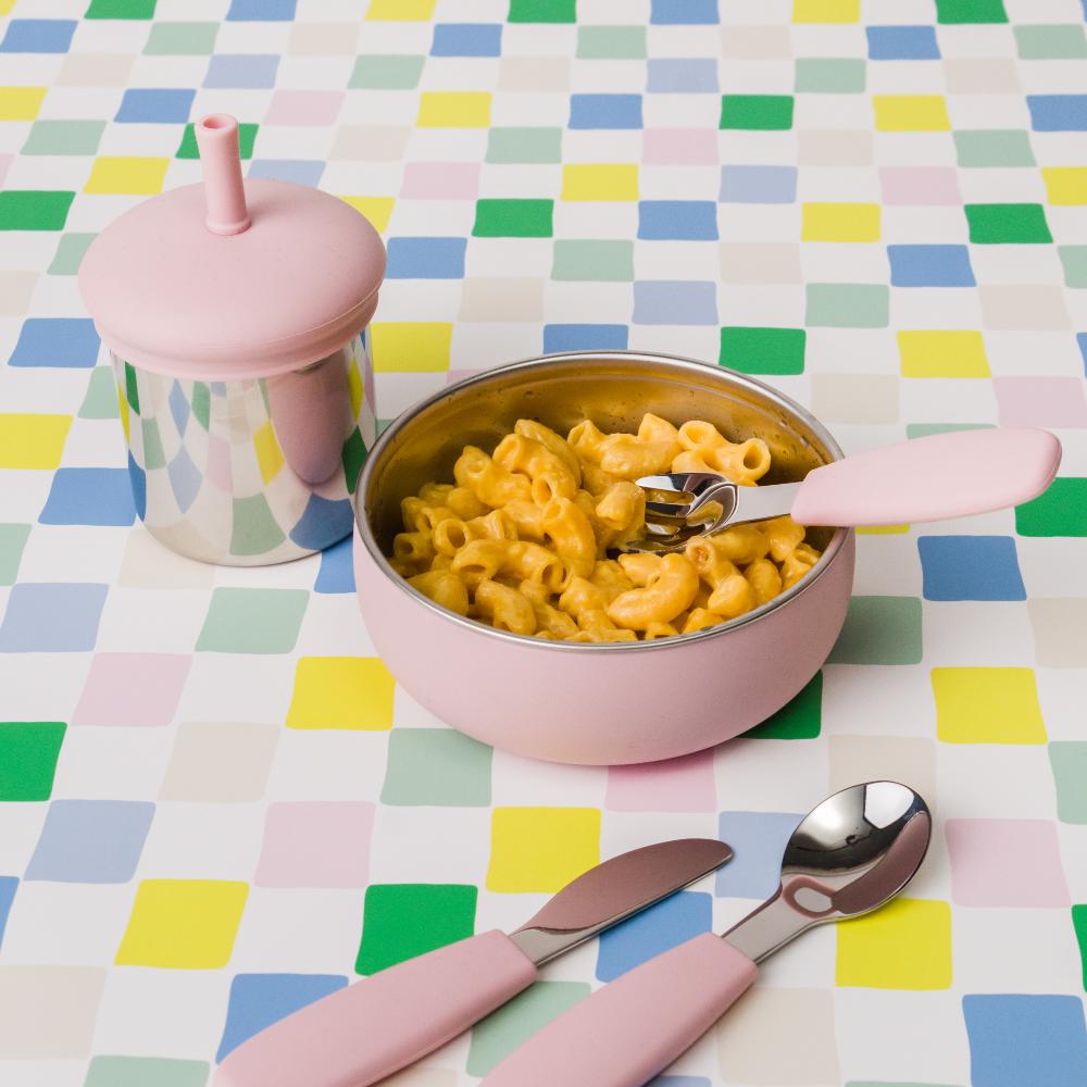 Pink stainless steel bowl with macaroni and cheese on a colorful checkered tablecloth, with pink utensils and a pink stainless steel cup with lid.