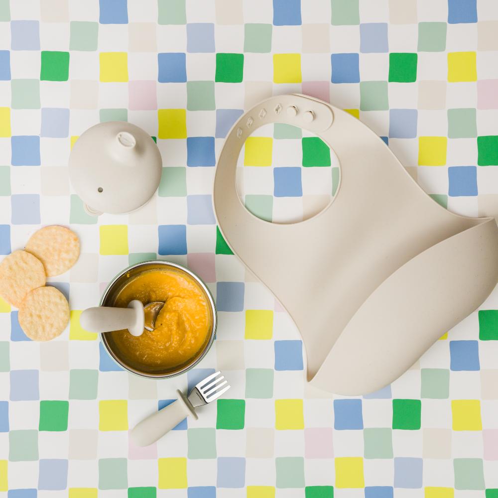 Nestling silicone baby bib, bowl with food, fork and spoon, and crackers on a checkered tablecloth