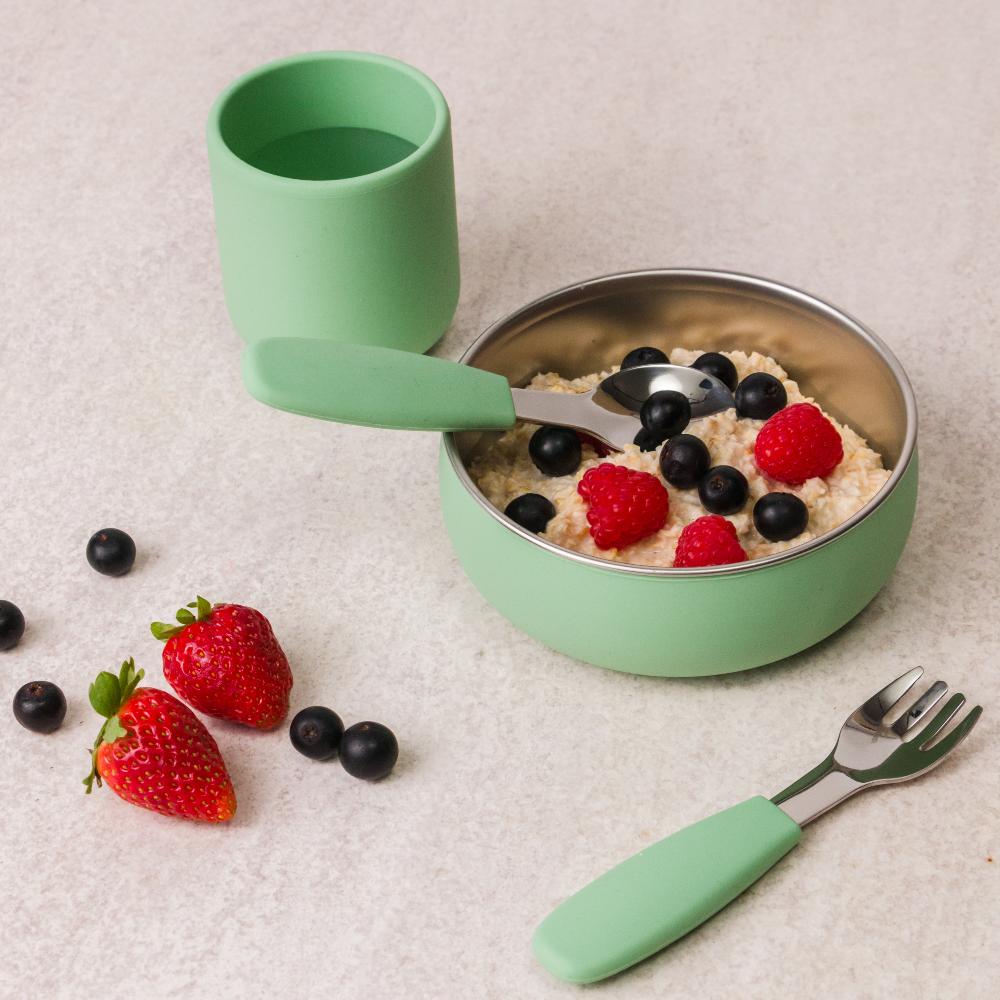Sage green children's bowl with spoon, fork, and silicone cup on a light surface with berries around.