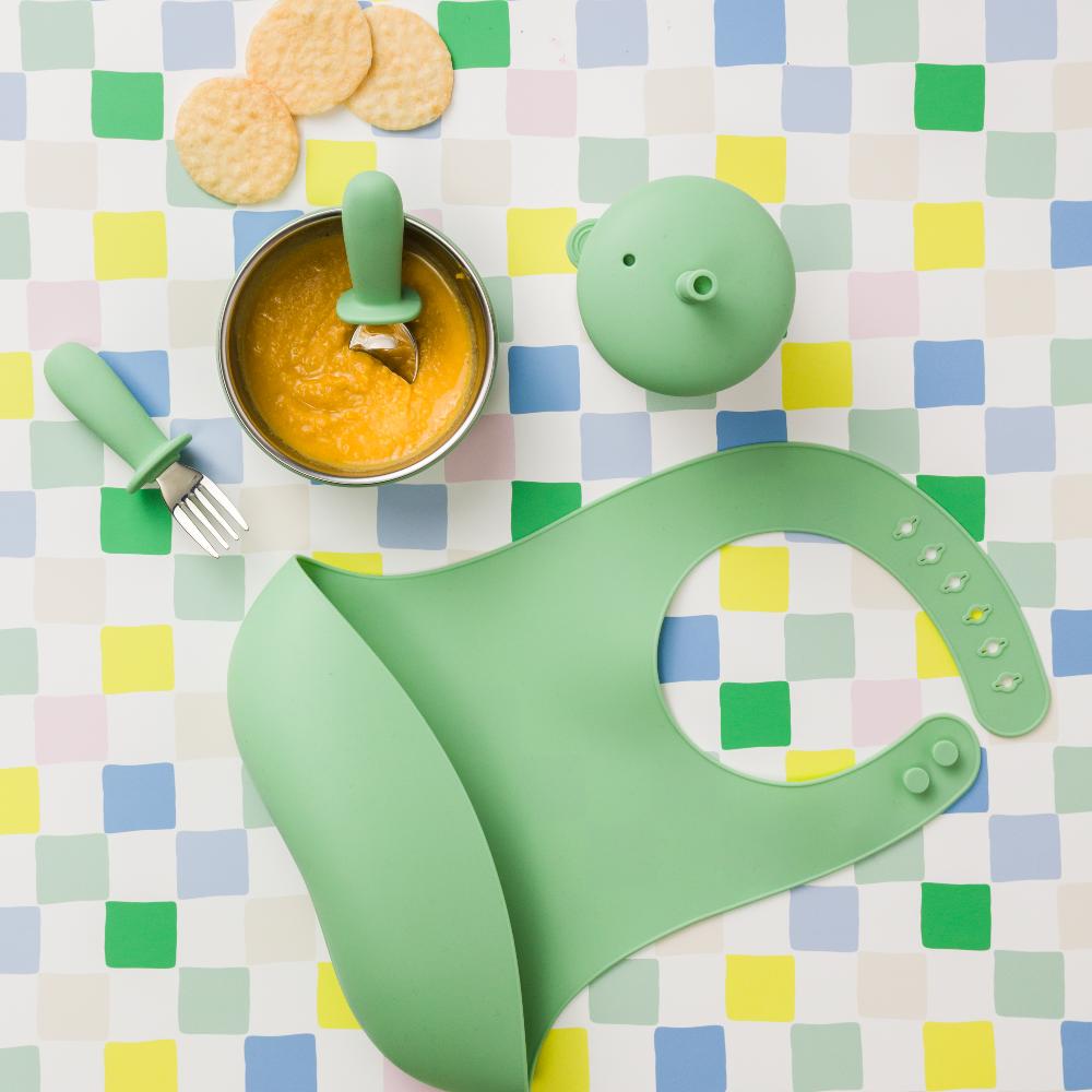 Silicone toddler bib with green feeding set with bowl, spoon and fork on a checkered tablecloth.