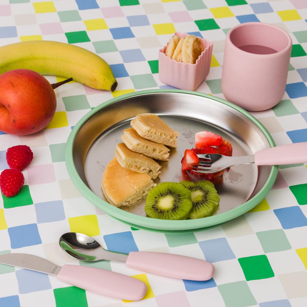 Toddler meal set with silicone training cup and stainless steel plate with pancakes, fruit, and utensils on a colorful checkered tablecloth.