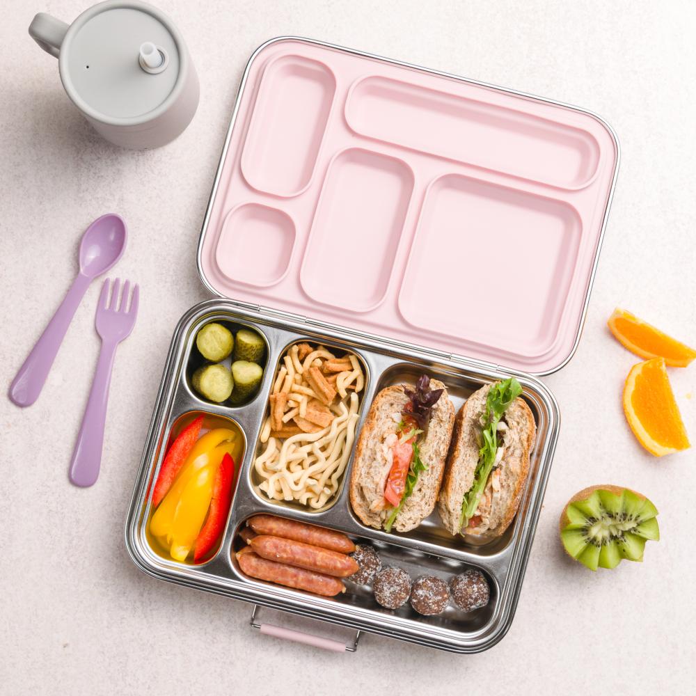 Bento box with compartments filled with food on a light surface with a teapot and fruit.