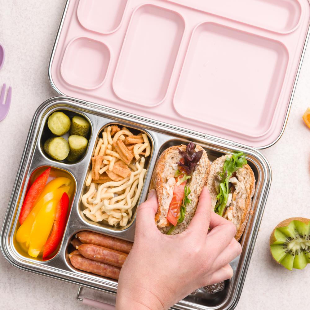 Pink bento box with compartments containing a sandwich, noodles, pickles, and vegetables on a light surface.