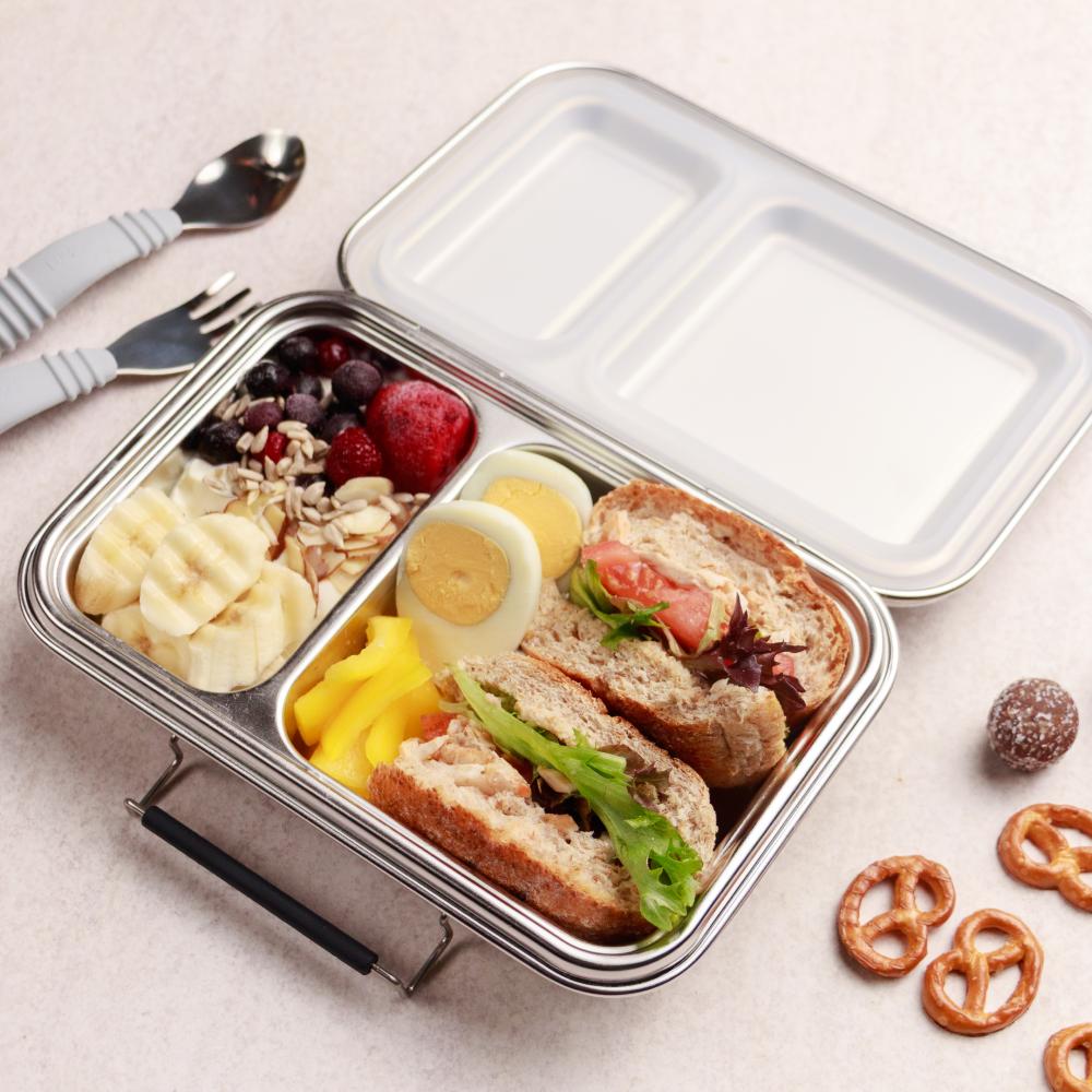 Metal lunch box with compartments containing a sandwich, fruit, and other snacks on a light surface.