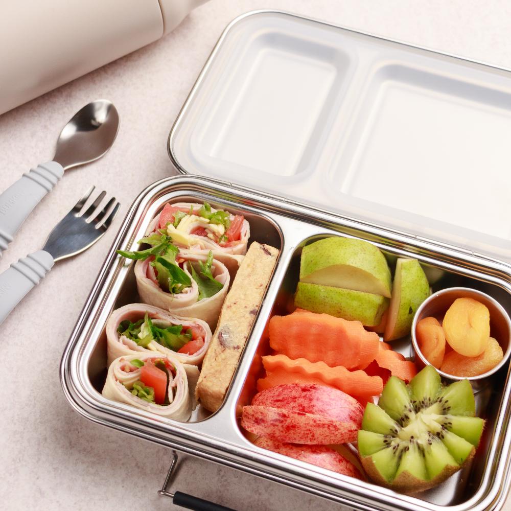 Metal lunch box with compartments filled with various food items including fruit and vegetables, on a light surface.