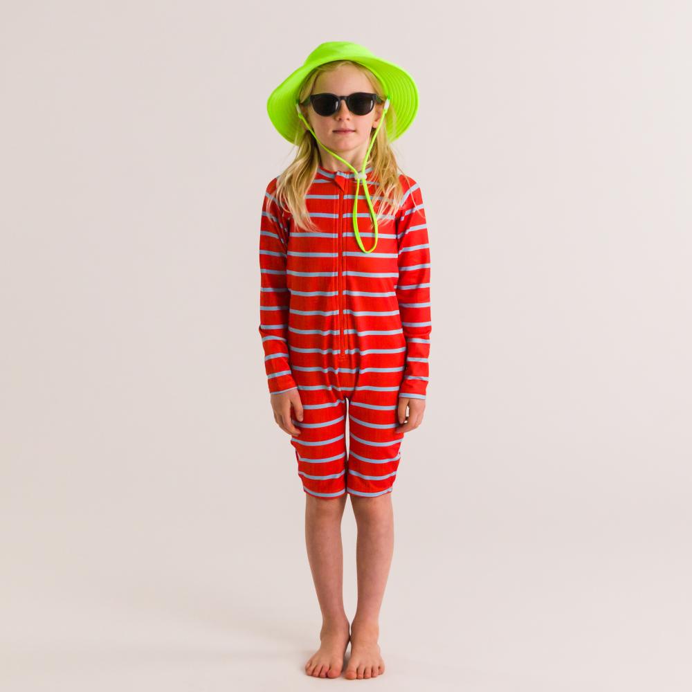 Child wearing a red and white striped swim suit with a green hat and sunglasses on a plain background